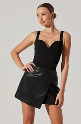 A woman is wearing a fitted black sleeveless top with thick straps and a sweetheart neckline, paired with a high-waisted black leather skirt that features a side wrap design, a button detail, and a slit. She accessorizes with gold hoop earrings and rings.